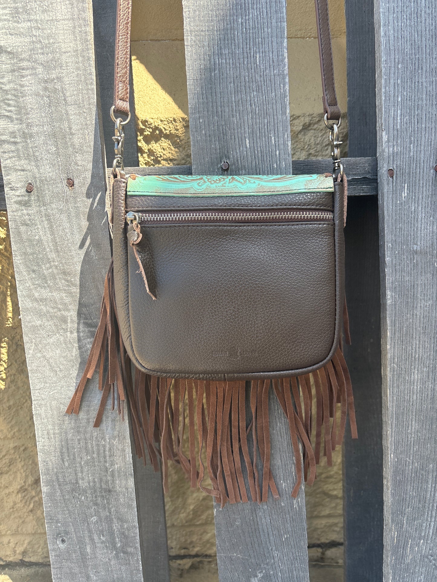 This distressed Turquoise tooled leather crossbody bag has brown fringe, brown strap, and a brown back. It features a whip stitch border around the tooled leather flap. Rear view showing zip close pocket. 