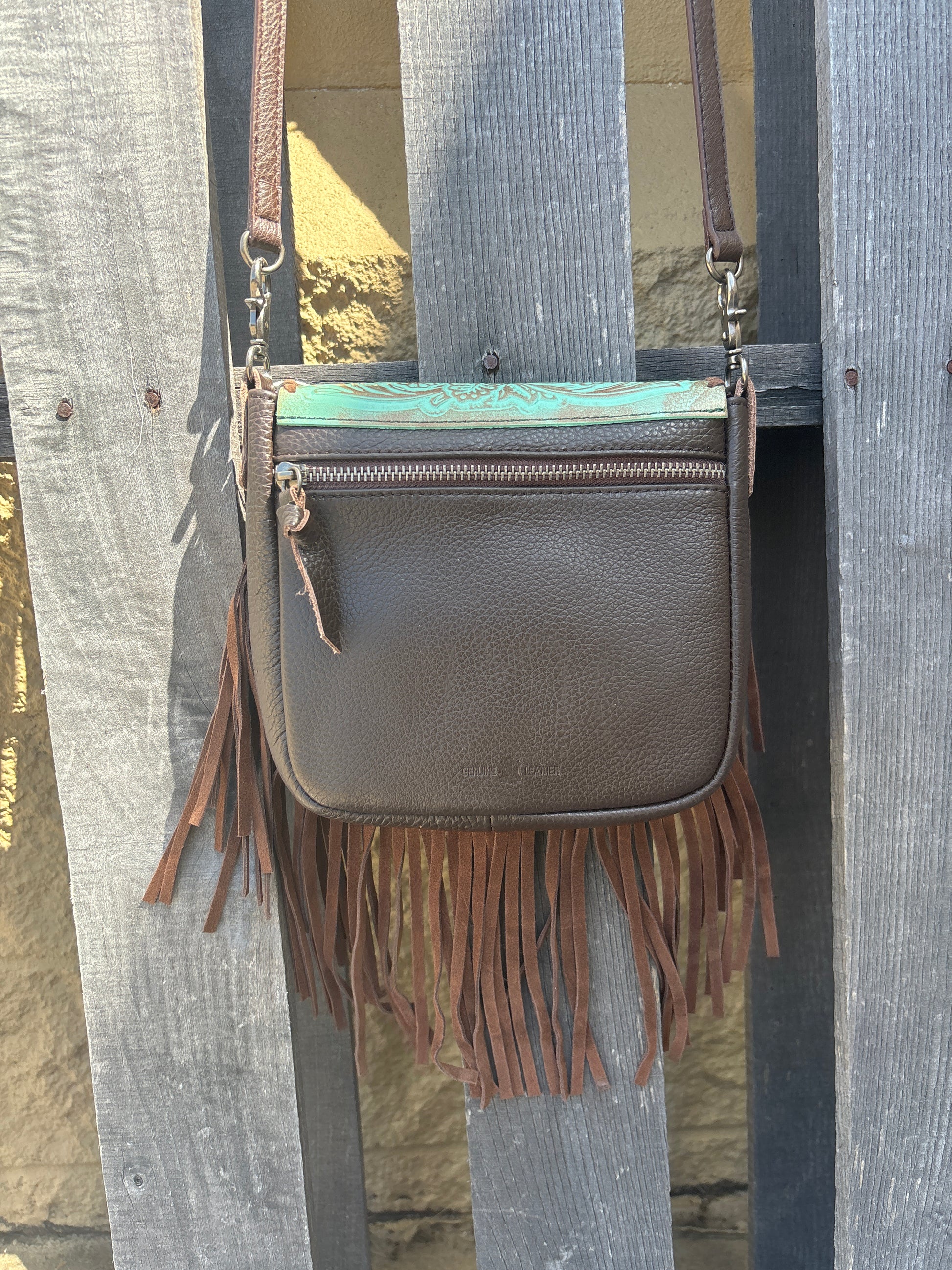 This distressed Turquoise tooled leather crossbody bag has brown fringe, brown strap, and a brown back. It features a whip stitch border around the tooled leather flap. Rear view showing zip close pocket. 