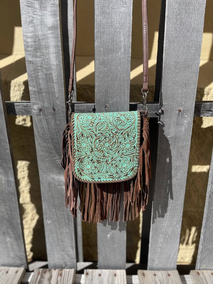 This distressed Turquoise tooled leather crossbody bag has brown fringe, brown strap, and a brown back. It features a whip stitch border around the tooled leather flap. Front View