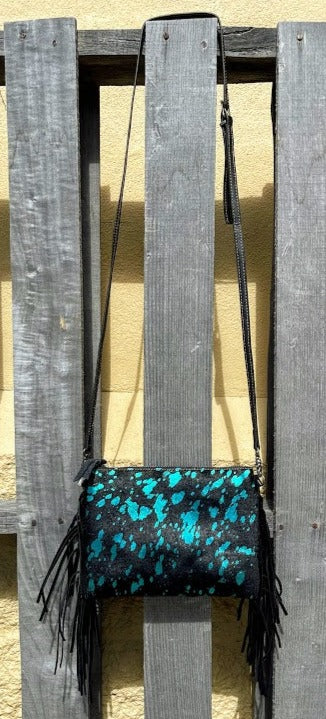 A small crossbody bag with turquoise and black cowhide and fringe hanging against a wooden background.