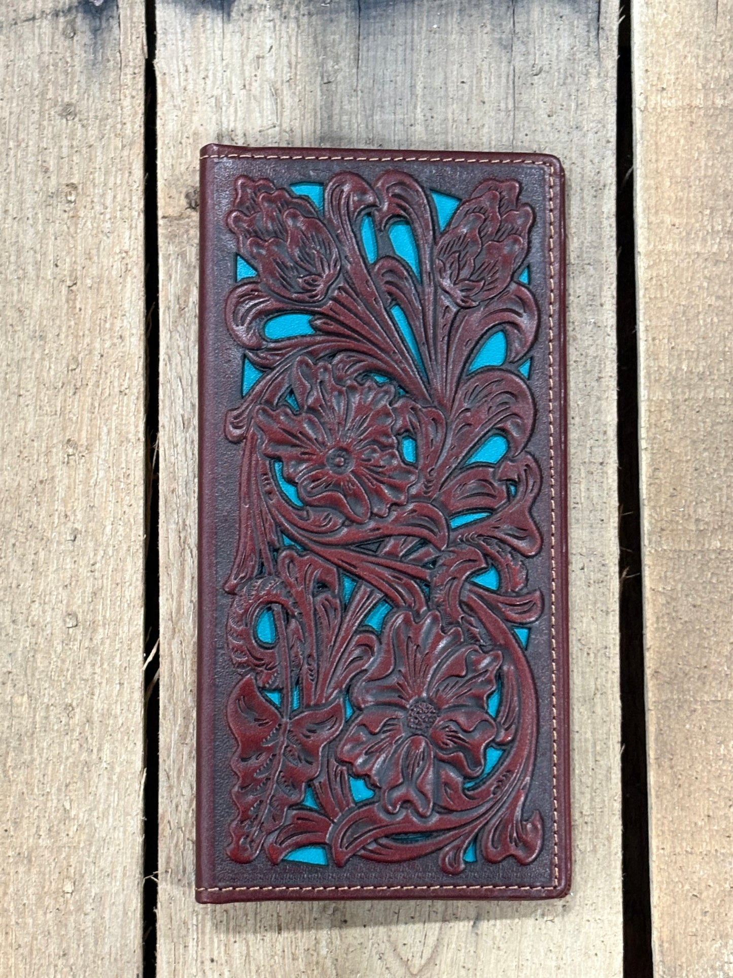 Brown leather wallet with intricate floral patterns on a striped fabric surface with a wooden wheel in the background.