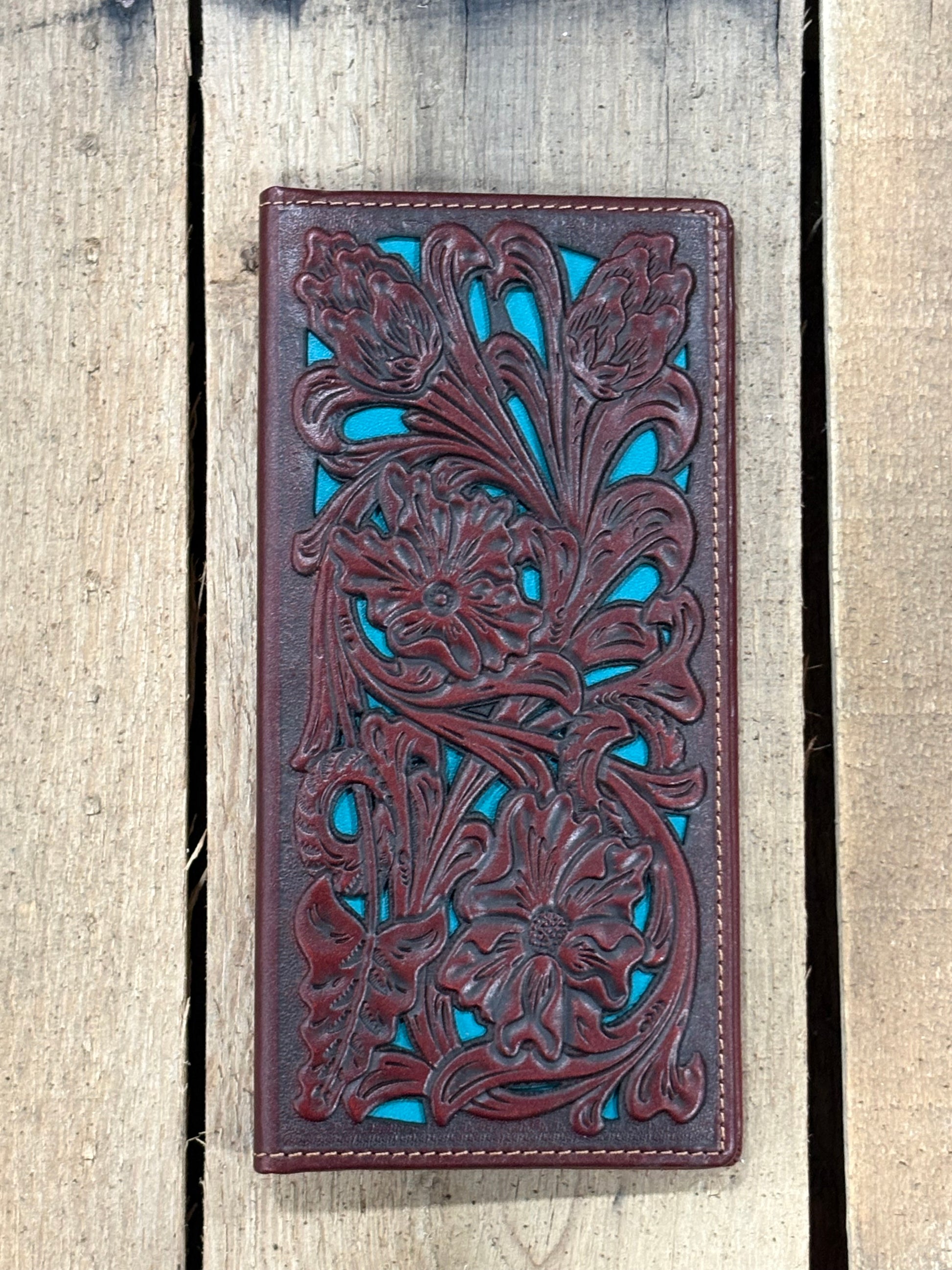 Brown leather wallet with intricate floral patterns on a striped fabric surface with a wooden wheel in the background.