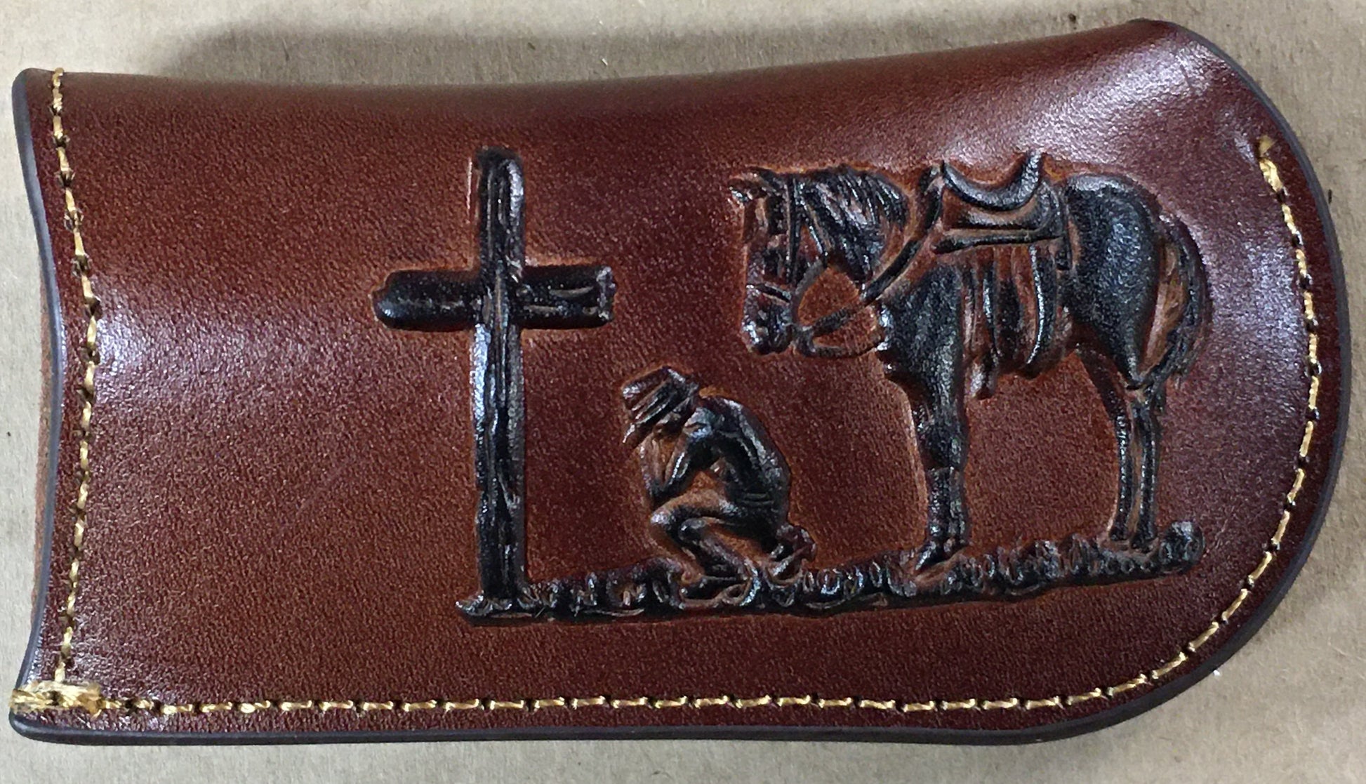 Brown leather case with metal cross, horse, and person kneeling design on a beige background