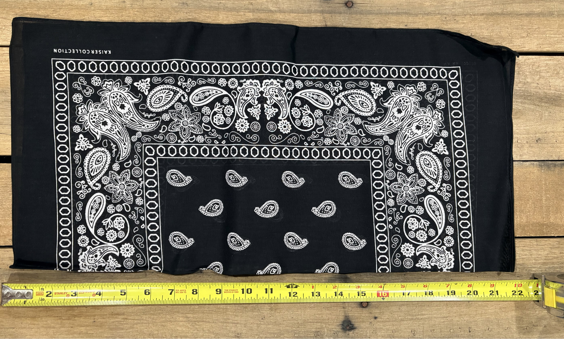 Black bandana with white paisley patterns on a wooden surface, measured by a yellow tape measure.