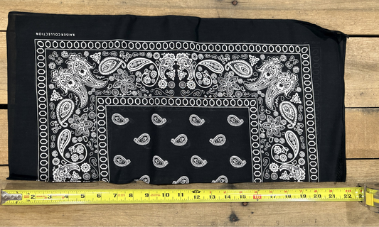 Black bandana with white paisley patterns on a wooden surface, measured by a yellow tape measure.