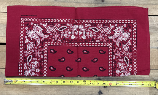 Burgundy bandana with white paisley patterns on a wooden surface, measured by a yellow tape measure.