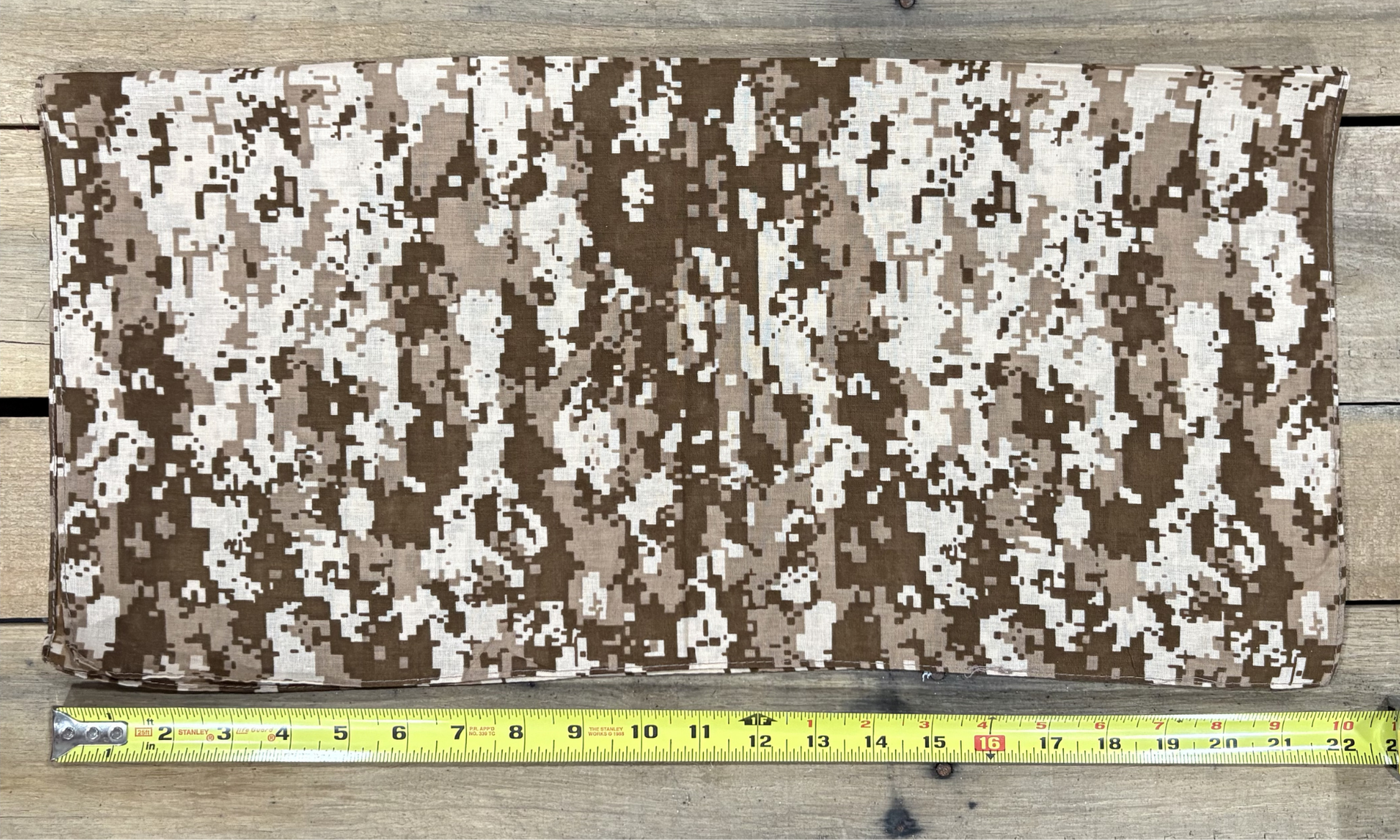Digital Camo Brown bandana with white paisley patterns on a wooden surface, measured by a yellow tape measure.