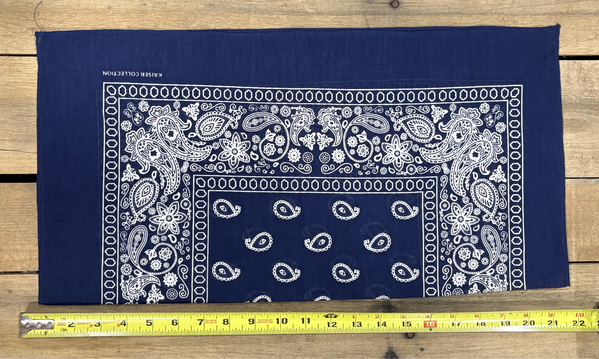 Navy bandana with white paisley patterns on a wooden surface, measured by a yellow tape measure.