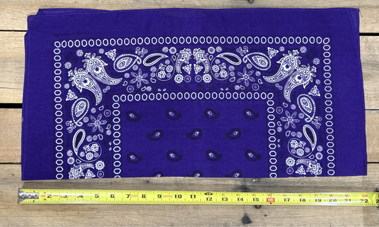 Purple bandana with white paisley patterns on a wooden surface, measured by a yellow tape measure.