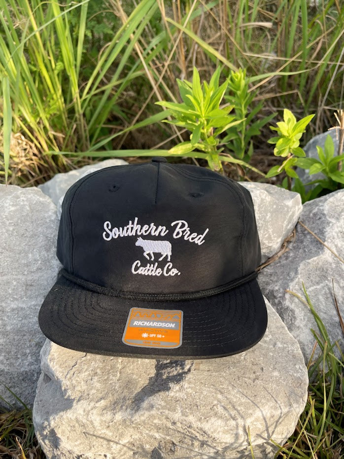 Black cap with 'Southern Bred Cattle Co.' text on a rock with grass in the background