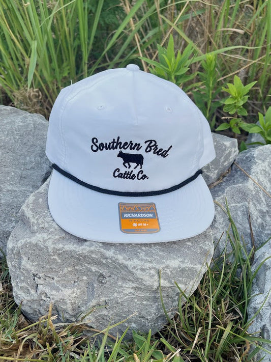 White cap with 'Southern Bred Cattle Co.' logo on a rock with grass and rocks in the background
