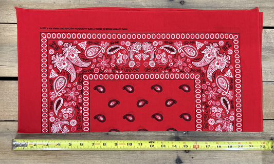 Red bandana with white paisley patterns on a wooden surface, measured by a yellow tape measure.