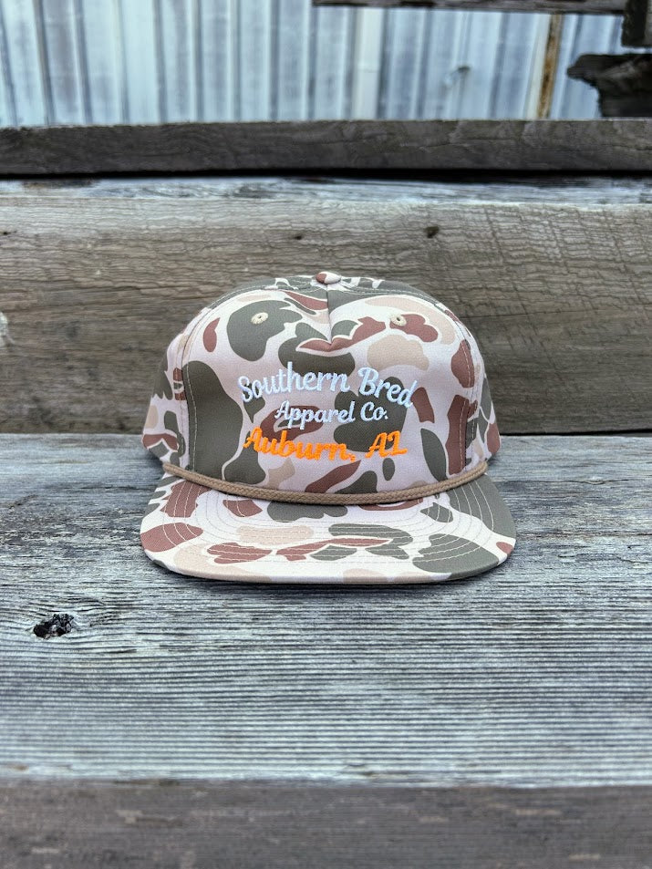 Camouflage-patterned cap with 'Southern Bred Apparel Co.' text on a wooden surface.