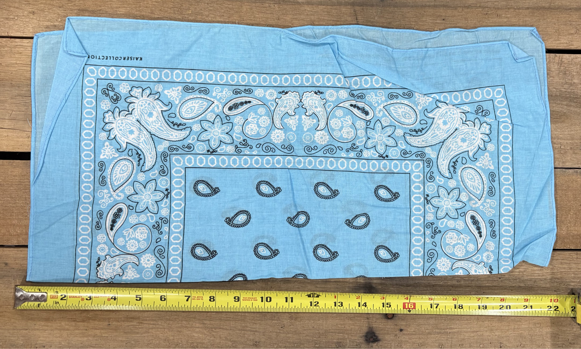 Sky Blue bandana with white paisley patterns on a wooden surface, measured by a yellow tape measure.